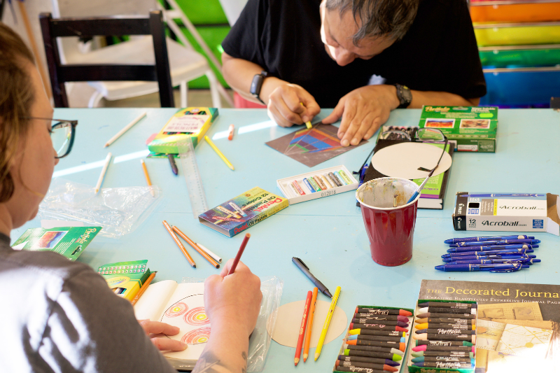 Individuals creating art at a table with various art supplies and utensils