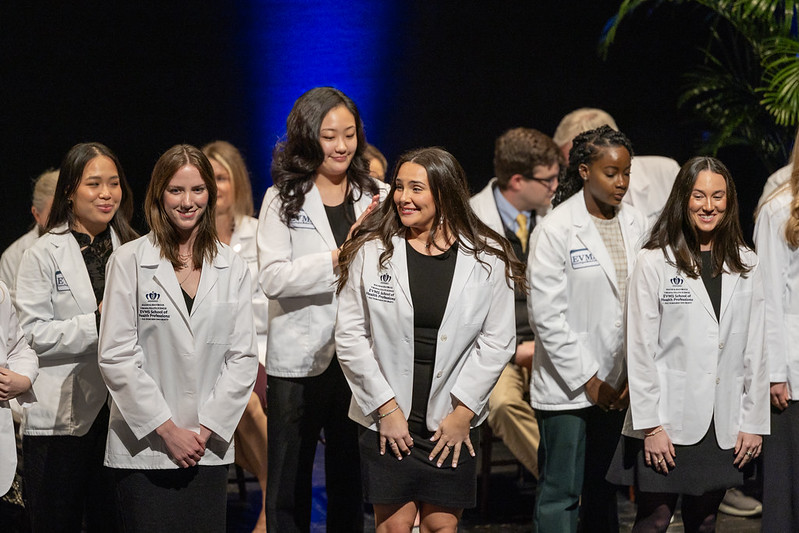 Students wearing white coats smile on stage during the ceremony