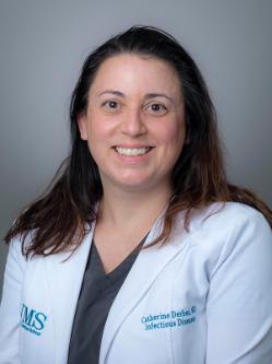 Dr. Catherine Derber is a woman with long dark brown hair, wears a white doctor coat and smiles at the camera