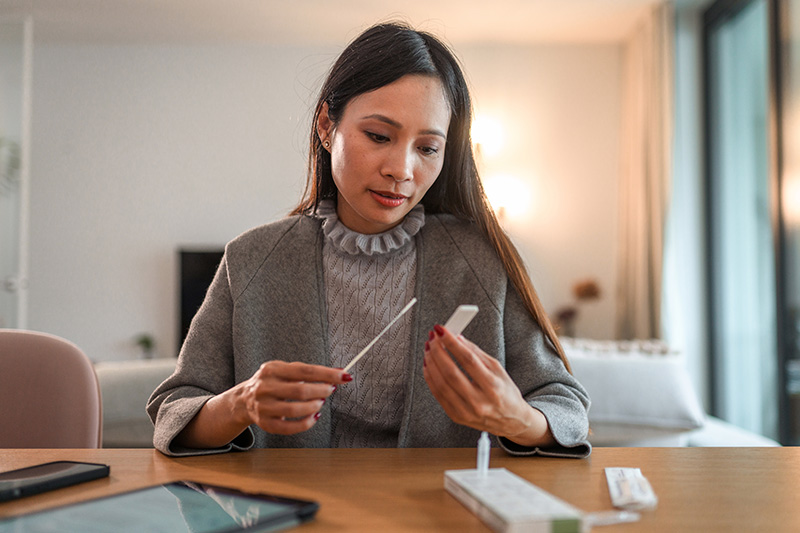 A business woman dressed in a sweater and blazer sits at a table at home holding a covid-19 rapid test awaiting results.