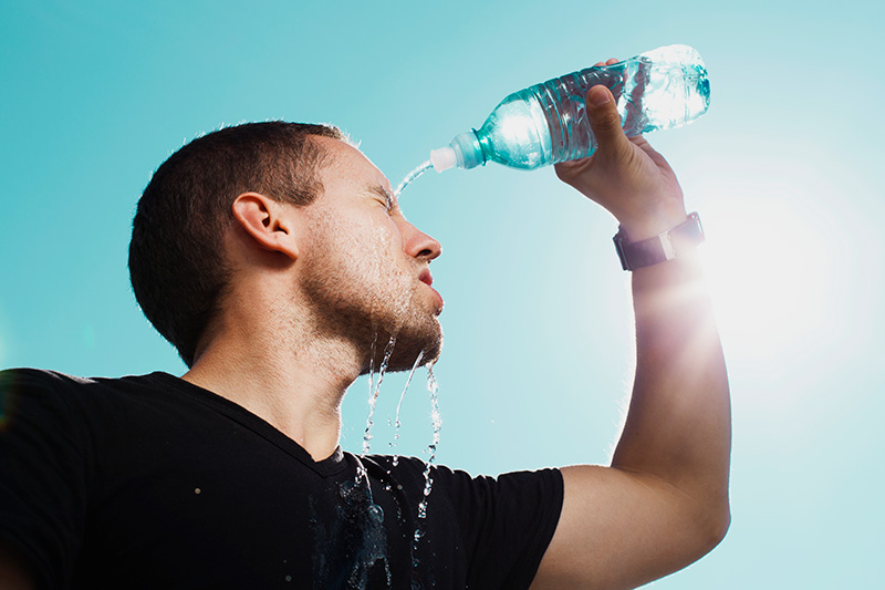 A man wearing a black t-shirt is pouring water from a bottle over his head while standing in the hot sun.