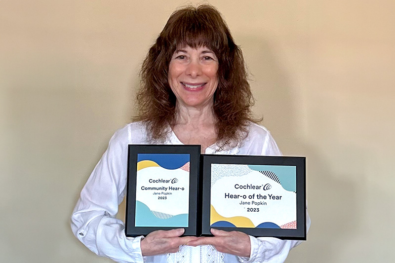 Jane has long curly brown hair, is wearing a white jacket and holds two award certificates while smiling at the camera