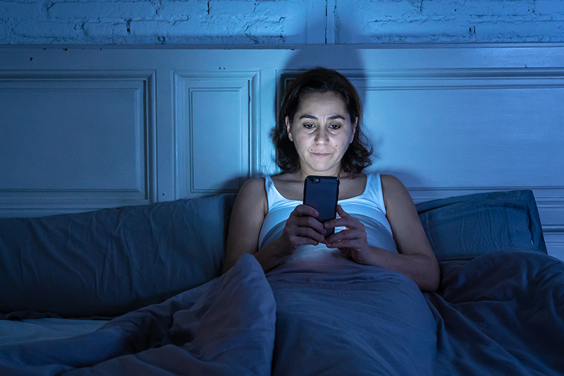 A woman sits up in bed in a dark room, looking at her cell phone.