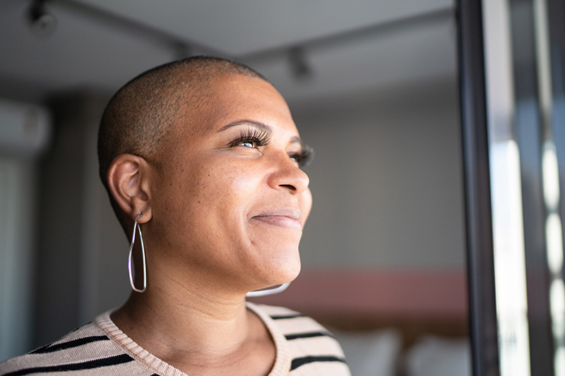 A woman with buzzed hair wearing a striped blouse smiles while looking out of a window.