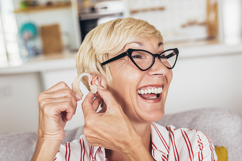 A senior woman with short blond hair and retro black eyeglasses puts on a hearing aid while smiling.