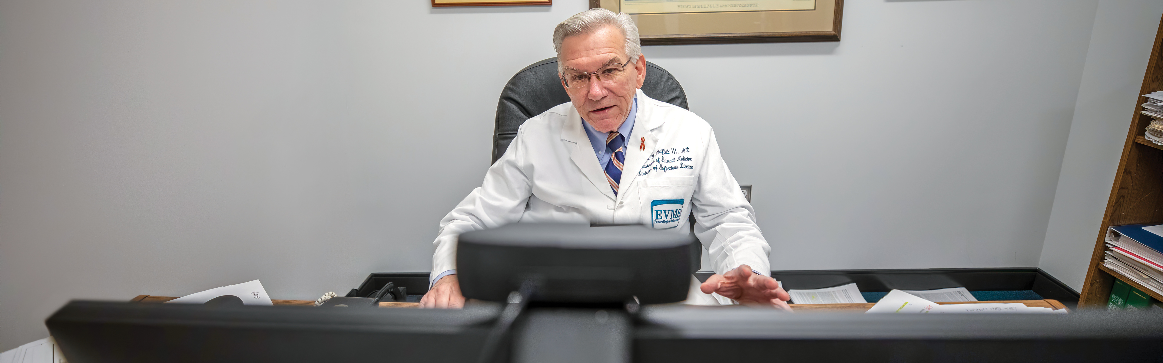 •	Dr. Edward Oldfield sits in front of his computer at his office desk while on a video conference call. There is a web cam attached to the top of his computer monitor.