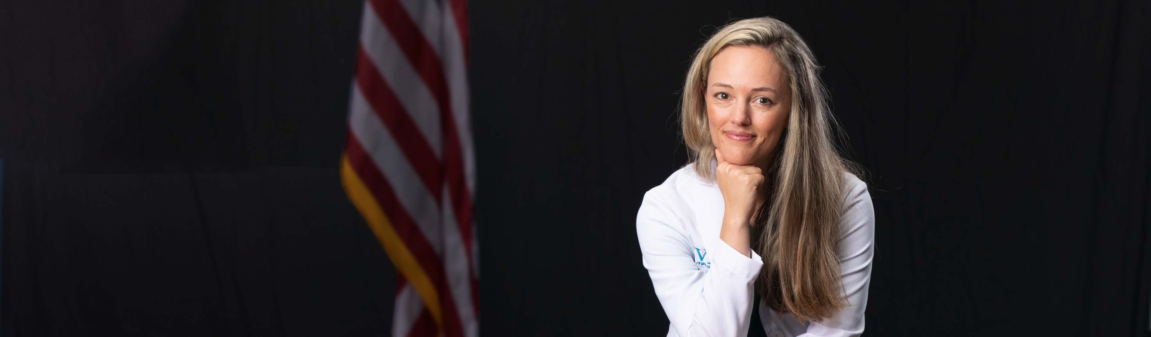 Dr. Brooke Hooper smiles at the camera and stands confidently in her white coat with the American flag out of focus behind her.