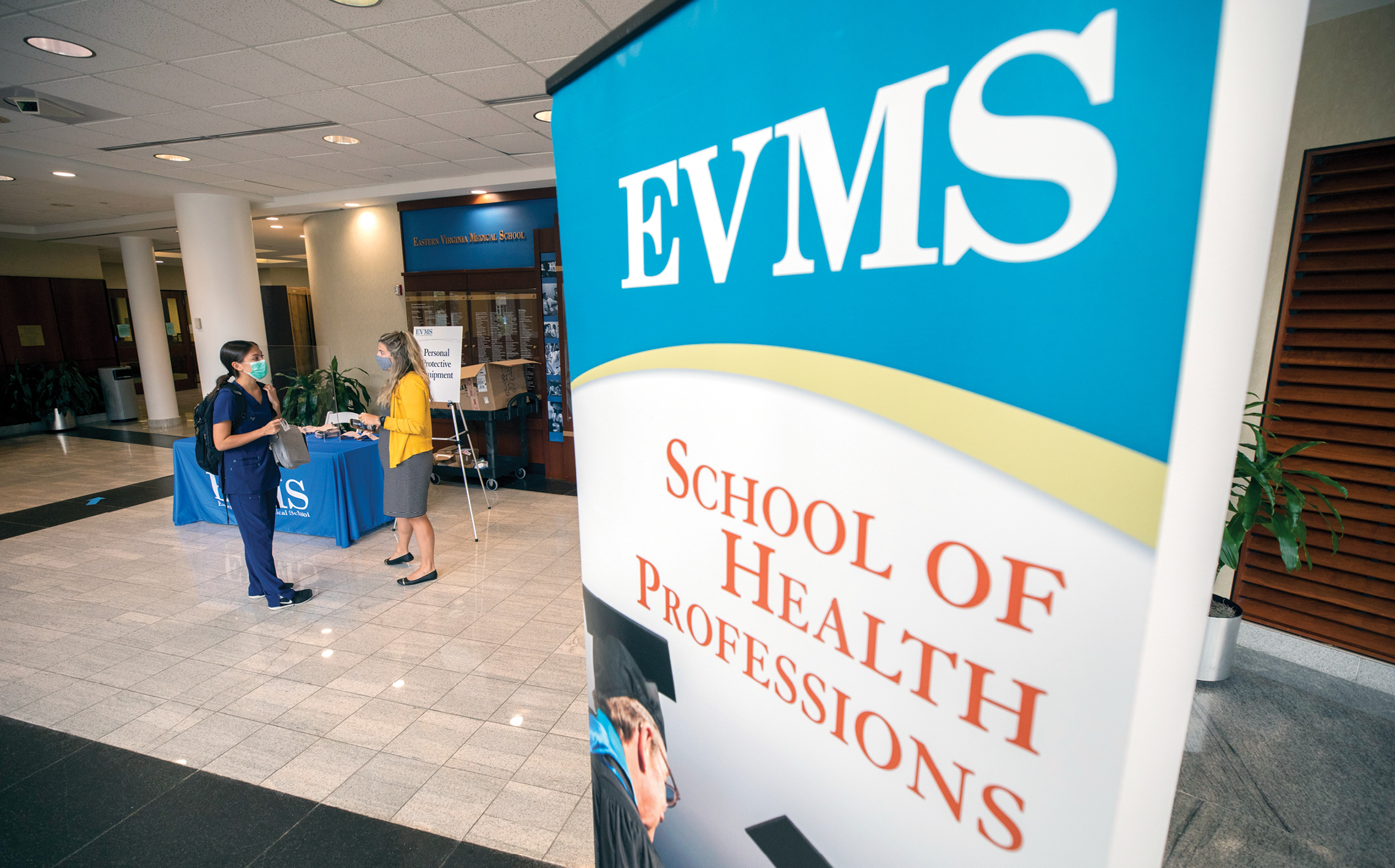 A student wearing a mask talks to faculty member while picking up her orientation materials at Lester Hall. A pull up banner in the right frame of the photo reads 