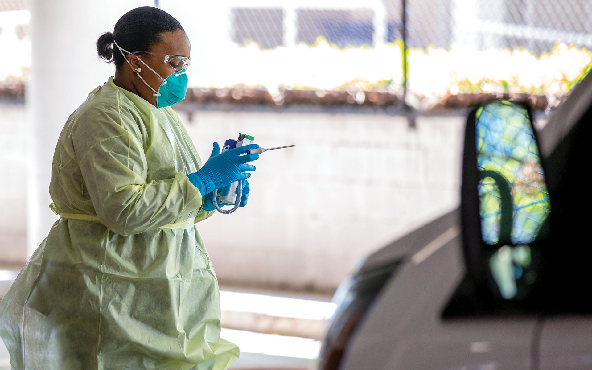 Adrienne Davis holds a thermometer before taking a patient's temperature at the COVID-19 drive-thru. She is wearing PPE including a face mask, goggles, gloves and a medical gown. The patient's car is in the right of the frame of the photo.