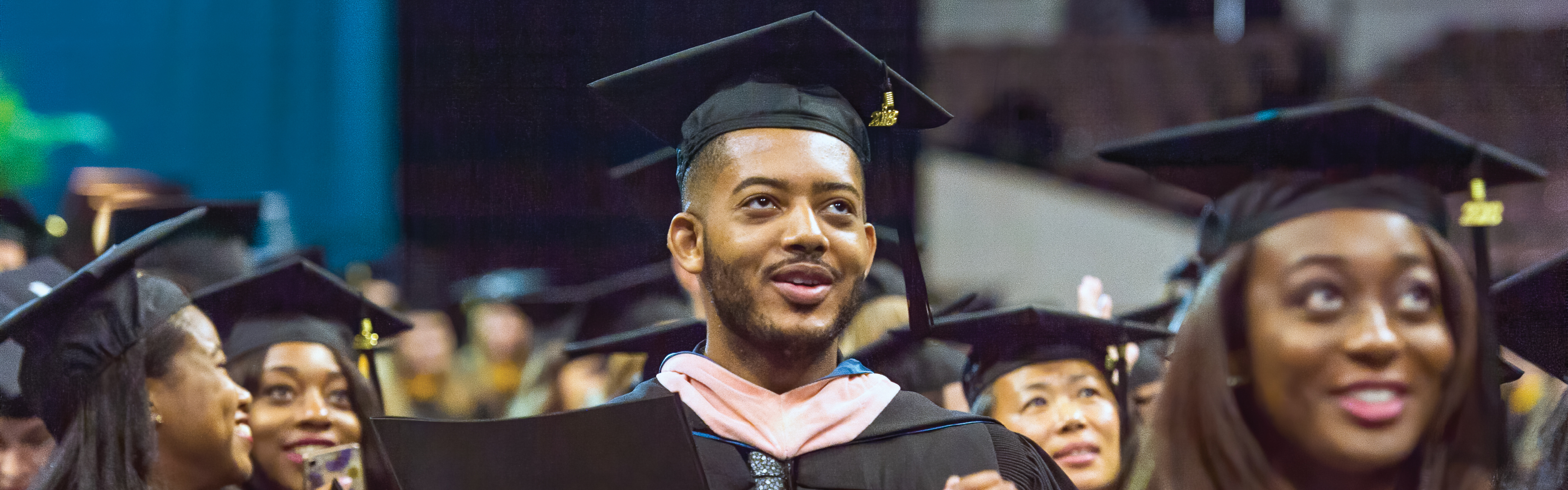 A group of diverse students at an EVMS Commencement ceremony. The figure in focus is a Black male graduate in cap and gown with a hopeful expression.