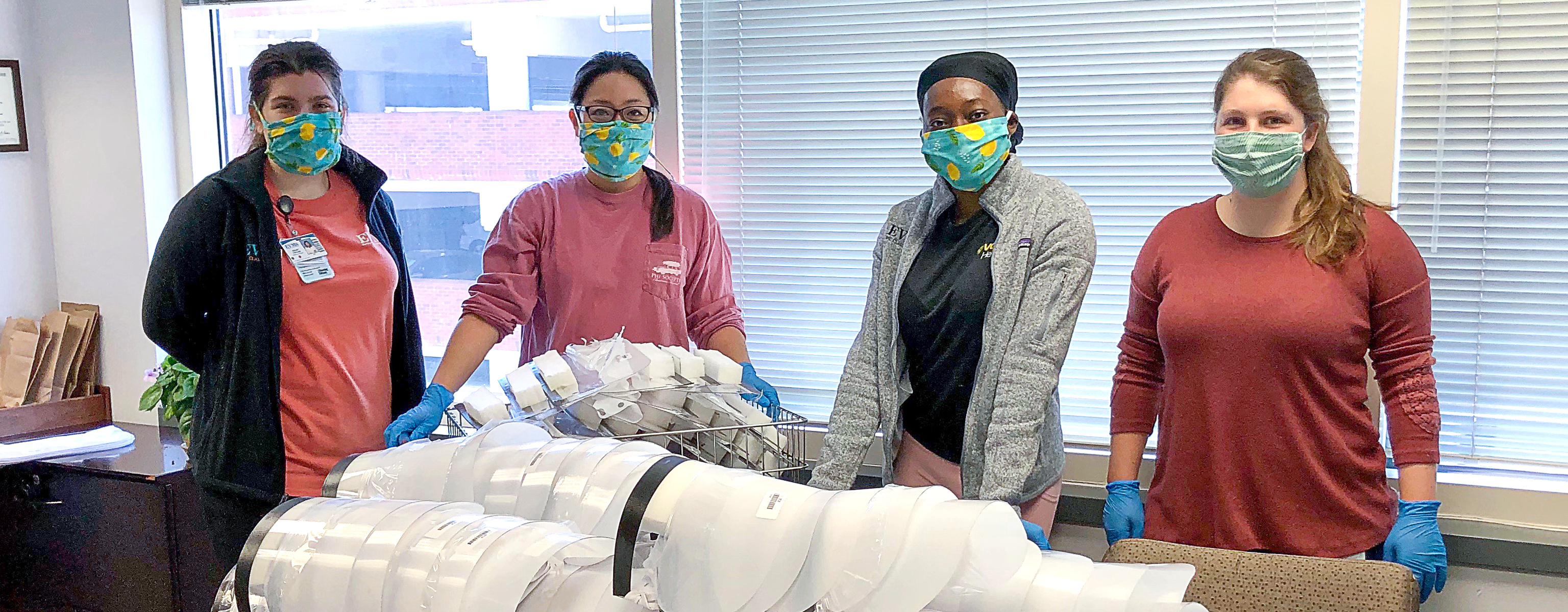 Four young women stand behind a desk covered in assembled face shields and face shield parts.