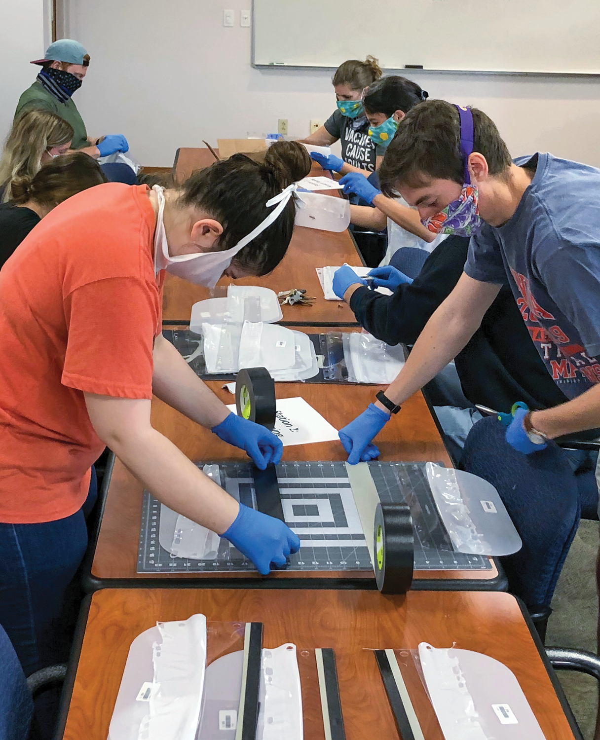 A female and male student in the foreground wear face covering and protective gloves as they use duct tape to cover the holes on the plastic coverings used to make the face shields.